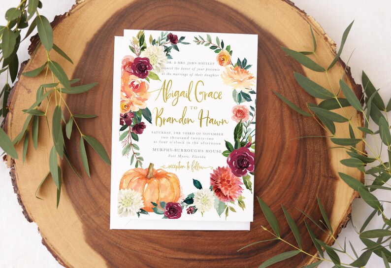 Op de afbeelding: Een huwelijksuitnodiging met een aquarelbloemenkrans, een pompoen en de tekst "Abigail Grace to Branden Hawn" in gouden letters. De uitnodiging ligt op een houten achtergrond.