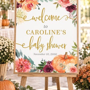 May include: A rectangular sign in a wooden frame featuring the text "welcome to CAROLINE'S baby shower November 10, 2030" in gold and black lettering, decorated with watercolor illustrations of orange pumpkins and various red and white flowers.