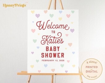 May include: A white sign on a wooden easel with the text "Welcome to Katie's Baby Shower February 12, 2026." The sign is decorated with pastel-colored hearts. A circular graphic indicates the sign is available in 4 sizes, printed or digital.