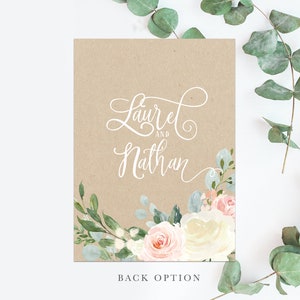 May include: A kraft paper wedding invitation with a floral design. The invitation features a white floral wreath with pink and white roses and green leaves. The text "Laurel and Nathan" is written in white cursive script.