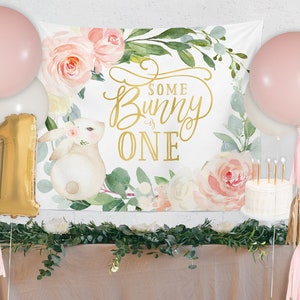 以下が含まれることがあります： 「Some Bunny is ONE」と金色の文字で書かれたバナーがある誕生日のパーティーシーン。バナーには水彩画のウサギと花のアクセントが描かれています。ピンクと白の風船、金の数字1の風船、ケーキが、お祝いの装飾を完成させています。