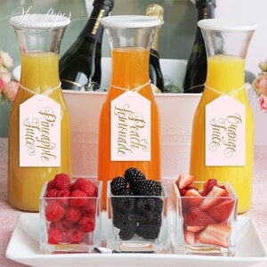 Może przedstawiać: Trzy szklane dzbanki wypełnione sokiem pomarańczowym, brzoskwiniowym i ananasowym, każdy z r&oacute;żową etykietą z napisem "Orange Juice", "Peach Lemonade" i "Pineapple Juice" złotym pismem. Dzbanki stoją na białej tacy z małymi szklanymi miseczkami z malinami, jeżynami i truskawkami.