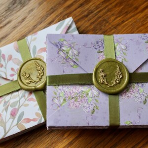 May include: Two sets of folded envelopes with floral patterns. The envelopes are tied with green ribbon and sealed with a wax seal featuring a gold wreath design.