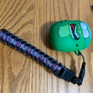 Puede incluir: Una funda verde para AirPod con forma de pepinillo, con una correa de paracord negra y un clip de plástico negro. La funda tiene una cara de dibujos animados con la boca muy abierta y ojos grandes.
