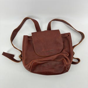 May include: A medium brown leather backpack featuring a fold-over flap closure, a drawstring top, and two adjustable shoulder straps. The bag has a smooth texture on the upper portion and a pebbled leather texture on the bottom panel.