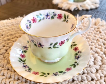 May include: A white teacup and saucer with a green and floral design. The cup has a gold rim and the saucer has a scalloped edge.