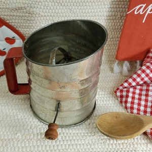 May include: A vintage silver-colored flour sifter with a red handle and a wooden knob. The sifter has measurements in cups and ounces.