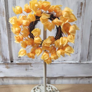 May include: A decorative wreath featuring vibrant yellow-orange flowers, arranged in a circular shape. The wreath is set against a white background and is supported by a white stand with a decorative base. The flowers have a delicate, papery texture.