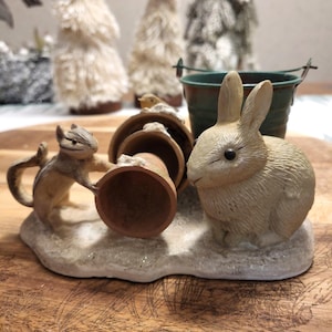 May include: A whimsical figurine featuring a chipmunk holding a terracotta pot, a seated rabbit, and a small green bucket. The scene is set on a snowy base, with miniature decorative trees in the background. The figurine is likely a decorative item.