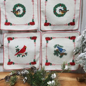 May include: Four square, off-white Christmas placemats with red trim and corner poinsettias. Each placemat features a different bird illustration: a cardinal, a blue jay, and two with a bird in a wreath. The placemats are approximately 12 inches square.