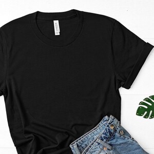 May include: A black t-shirt with a white tag that says "CANVAS." The t-shirt is laying on a white surface with a pair of blue jeans and a green leaf-shaped keychain.