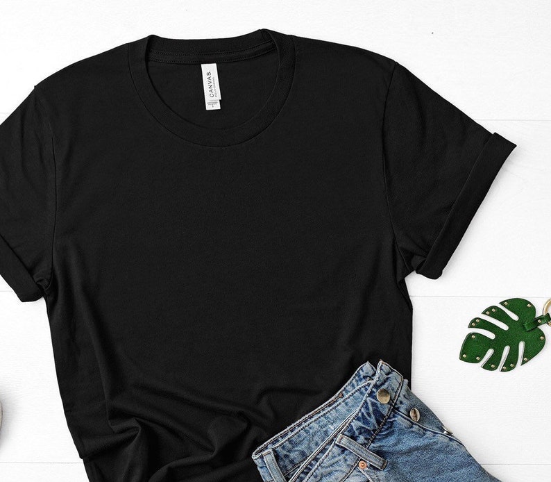 May include: A black t-shirt with a white tag that says "CANVAS." The t-shirt is laying on a white surface with a pair of blue jeans and a green leaf-shaped keychain.
