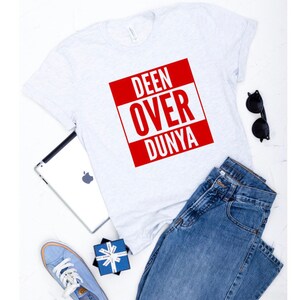 May include: A white t-shirt with a red square that says "DEEN OVER DUNYA" in white letters. The t-shirt is laying on a white surface with a pair of jeans, a tablet, sunglasses, and a blue gift box.