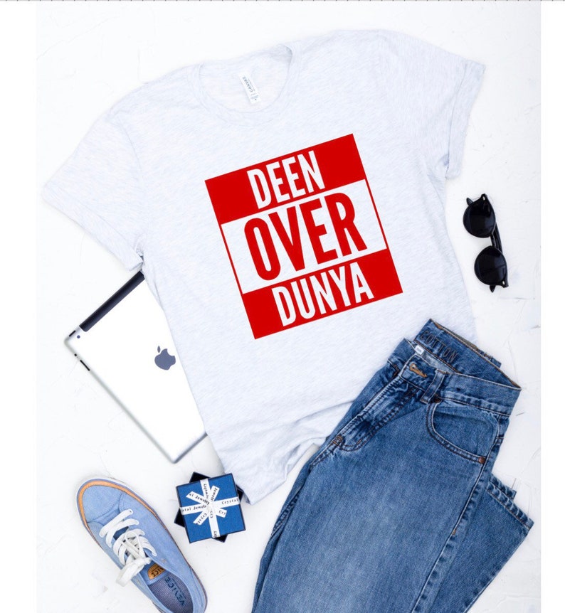 May include: A white t-shirt with a red square that says "DEEN OVER DUNYA" in white letters. The t-shirt is laying on a white surface with a pair of jeans, a tablet, sunglasses, and a blue gift box.