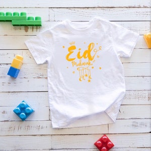 May include: White short-sleeved t-shirt with a yellow graphic design that says "Eid Mubarak" with a crescent moon and stars.