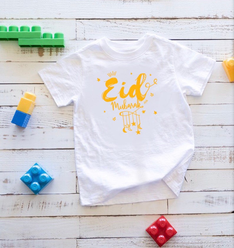 May include: White short-sleeved t-shirt with a yellow graphic design that says "Eid Mubarak" with a crescent moon and stars.