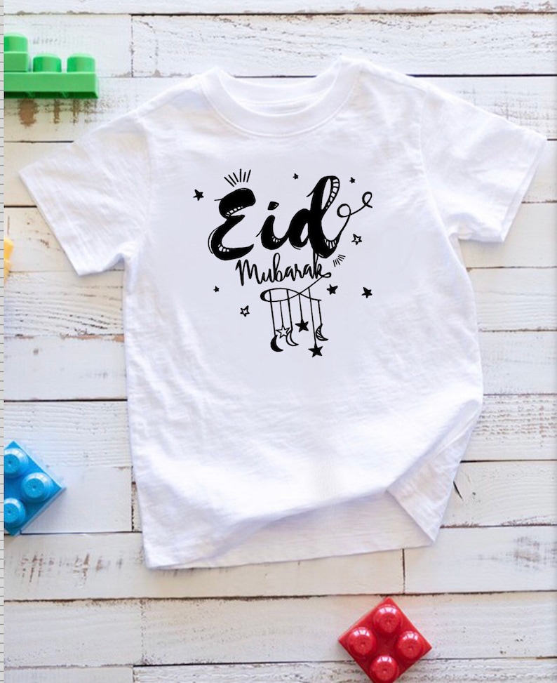 May include: White t-shirt with black text that says "Eid Mubarak" with a design of a crescent moon and stars hanging from a string.