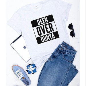 May include: A white t-shirt with a black square graphic that says "DEEN OVER DUNYA" in white letters. The shirt is paired with blue jeans and blue sneakers.