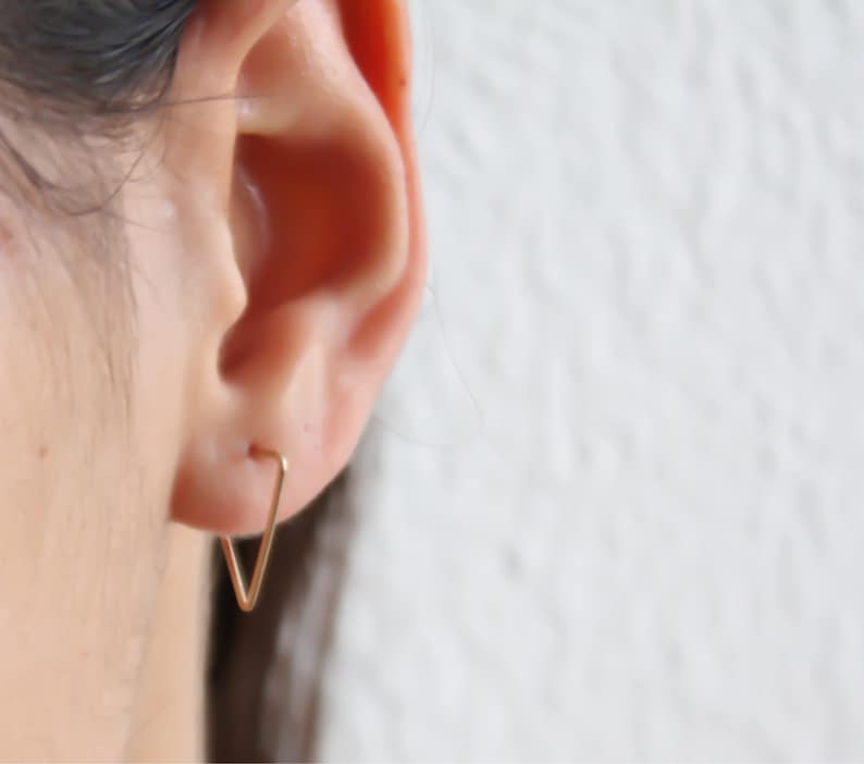 May include: A pair of gold triangle shaped earrings. The earrings are small and delicate, and they are worn in the wearer's earlobes.