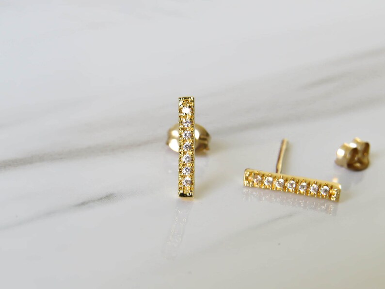 May include: A pair of gold bar earrings with small clear stones set in the bar. The earrings are on a white marble surface.