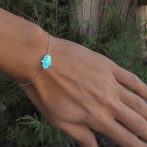 May include: A silver chain bracelet with a blue opal hamsa hand charm. The bracelet is on a person's wrist.