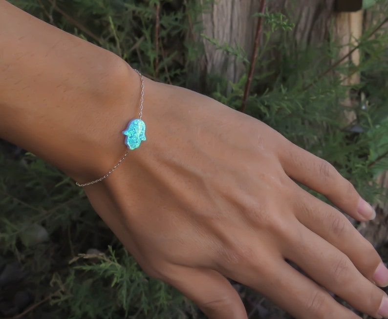 May include: A silver chain bracelet with a blue opal hamsa hand charm. The bracelet is on a person's wrist.