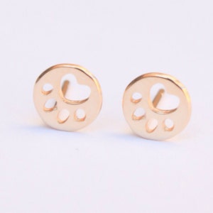 Op de afbeelding: Een paar gouden oorbel studs met een pootprint ontwerp. De pootprint is uit het midden van elke oorbel gesneden, wat een delicate en stijlvolle look cre&euml;ert.