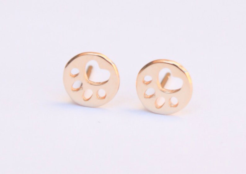 Op de afbeelding: Een paar gouden oorbel studs met een pootprint ontwerp. De pootprint is uit het midden van elke oorbel gesneden, wat een delicate en stijlvolle look cre&euml;ert.