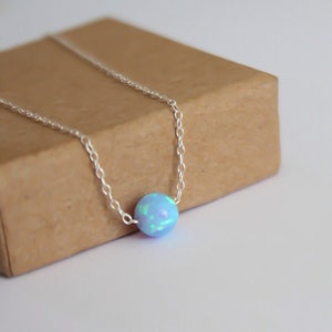 Puede incluir: Un delicado collar de cadena de plata con un solo colgante redondo de ópalo azul. El ópalo tiene una base azul claro con destellos iridiscentes verdes y azules.