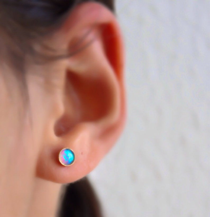 Puede incluir: Un par de peque&ntilde;os pendientes de bot&oacute;n de &oacute;palo redondo. Los pendientes est&aacute;n engastados en oro y tienen un brillo iridiscente azul y rosa.