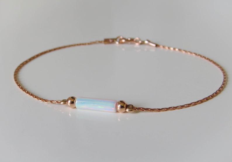 May include: A delicate rose gold chain bracelet with a white opal bar charm. The chain is a braided design and the charm is set between two small rose gold beads.