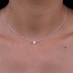 May include: A delicate silver necklace featuring a small, square, iridescent pendant. The pendant appears to be a cube shape, and it hangs from a thin, silver chain. The necklace is worn around the neck, showcasing a minimalist jewelry design.