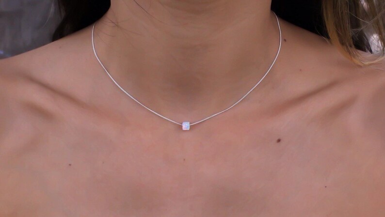 May include: A delicate silver necklace featuring a small, square, iridescent pendant. The pendant appears to be a cube shape, and it hangs from a thin, silver chain. The necklace is worn around the neck, showcasing a minimalist jewelry design.