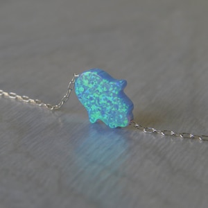 May include: A delicate silver chain necklace with a blue opal hamsa hand charm. The hamsa hand is a symbol of protection and good luck.