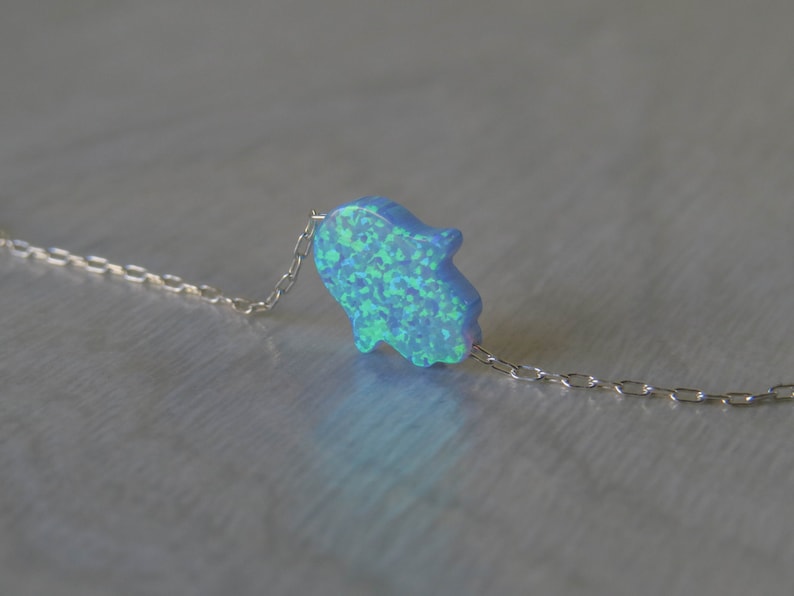 May include: A delicate silver chain necklace with a blue opal hamsa hand charm. The hamsa hand is a symbol of protection and good luck.