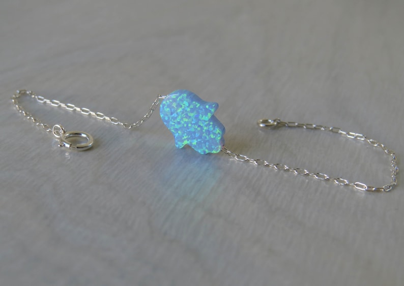 May include: A silver chain bracelet featuring a blue opal hamsa hand charm. The hamsa hand is a symbol of protection and good luck.