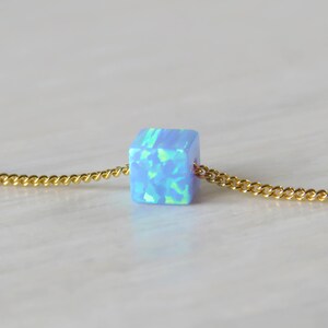 May include: A close-up of a necklace featuring a small, light blue cube pendant with a gold chain. The cube has a mosaic-like appearance with shades of blue and green. The gold chain is delicate and has a curb link design.