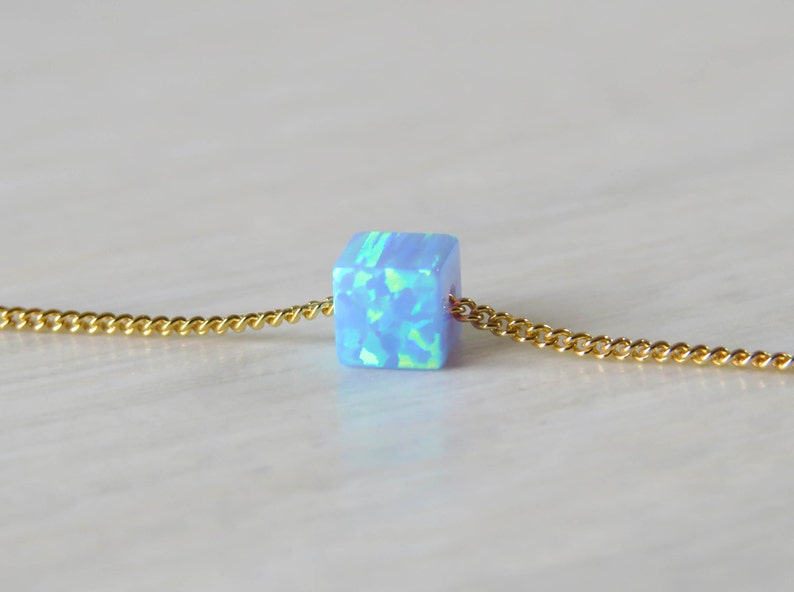 May include: A close-up of a necklace featuring a small, light blue cube pendant with a gold chain. The cube has a mosaic-like appearance with shades of blue and green. The gold chain is delicate and has a curb link design.