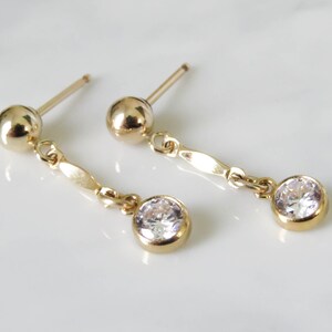 May include: A pair of gold earrings with a small round clear stone dangling from each earring.