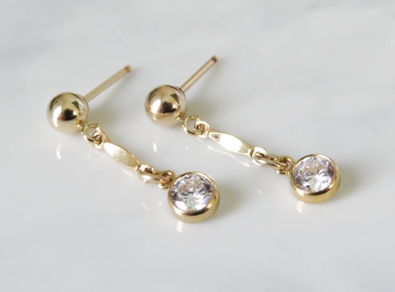May include: A pair of gold earrings with a small round clear stone dangling from each earring.