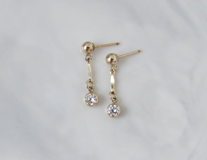 May include: A pair of gold dangle earrings with a small clear stone at the end of each earring.