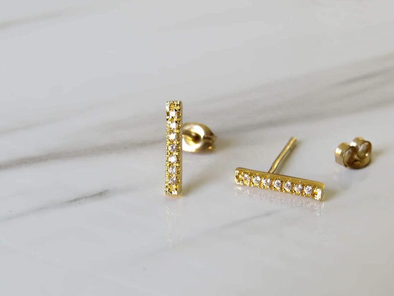 May include: A pair of gold bar earrings with small clear stones set in the metal. The earrings are on a white marble surface.