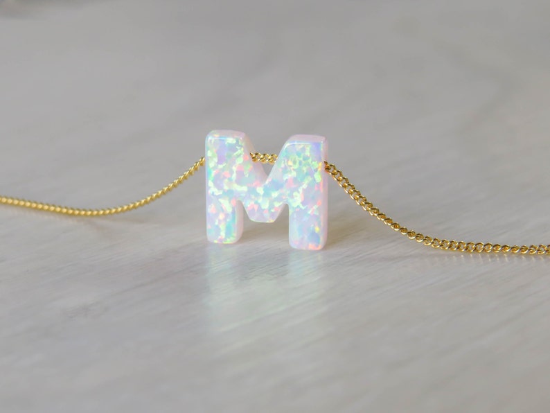 May include: A delicate gold chain necklace with a white opal letter M pendant. The opal has a rainbow iridescent sheen.