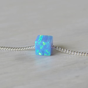 May include: A light blue opal cube pendant on a silver chain. The cube has a shimmering, iridescent quality with hints of green and blue. The chain is a delicate curb style, and the necklace is displayed on a light-colored surface.
