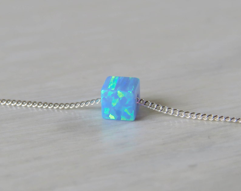 May include: A light blue opal cube pendant on a silver chain. The cube has a shimmering, iridescent quality with hints of green and blue. The chain is a delicate curb style, and the necklace is displayed on a light-colored surface.
