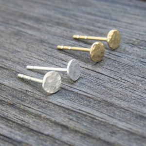 Op de afbeelding: Drie paar gehamerde metalen oorbellen, twee goud en een zilver, getoond op een houten oppervlak.
