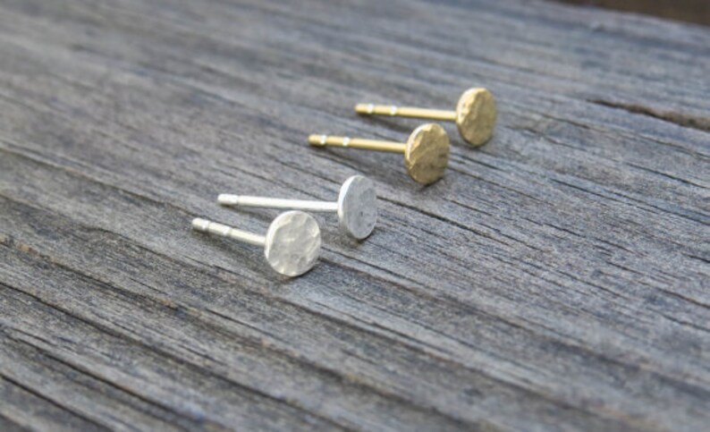 Op de afbeelding: Drie paar gehamerde metalen oorbellen, twee goud en een zilver, getoond op een houten oppervlak.