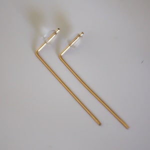 May include: A pair of gold-toned earrings with a simple, geometric design. Each earring features a straight, thin rod that bends at a right angle to form a short, horizontal bar. The earrings have clear plastic stoppers at the top.
