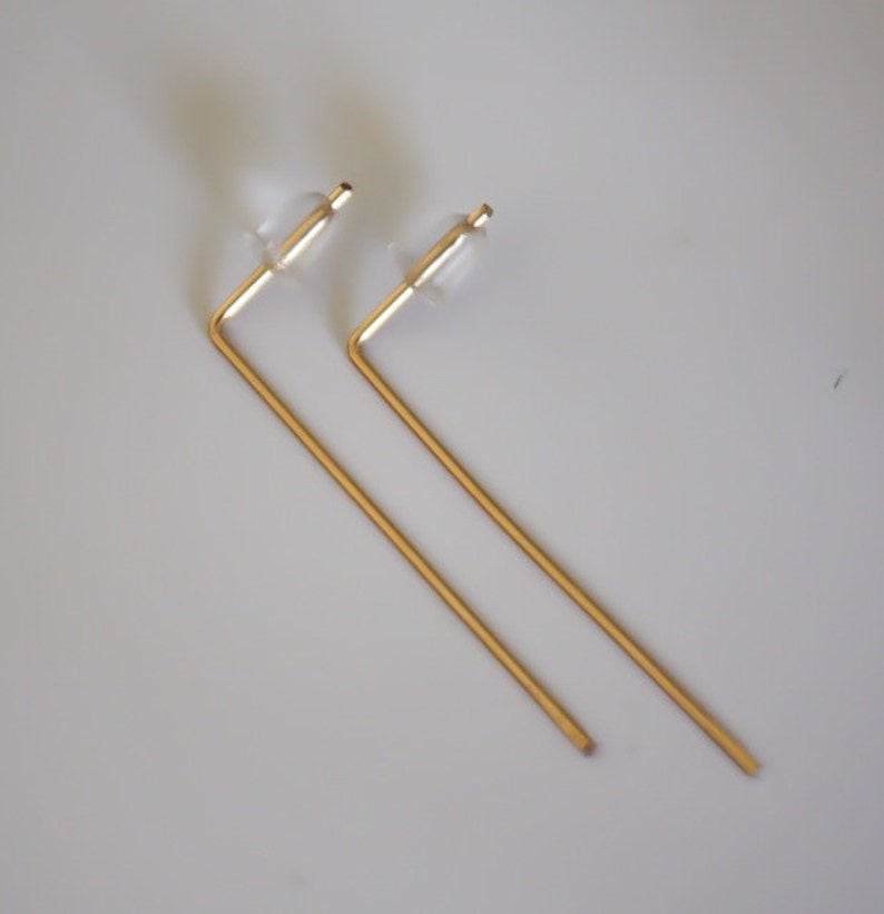May include: A pair of gold-toned earrings with a simple, geometric design. Each earring features a straight, thin rod that bends at a right angle to form a short, horizontal bar. The earrings have clear plastic stoppers at the top.