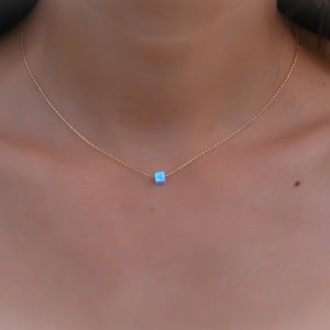 May include: A delicate gold necklace featuring a small, square, light blue pendant. The pendant is centered on a thin gold chain, creating a minimalist and elegant design. The necklace is shown against a skin tone background.